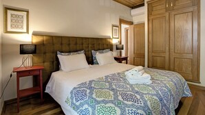 Un dormitorio con cabecera capitonada, una cama grande con ropa de cama estampada, un armario de madera y una mesita de noche con lámpara.