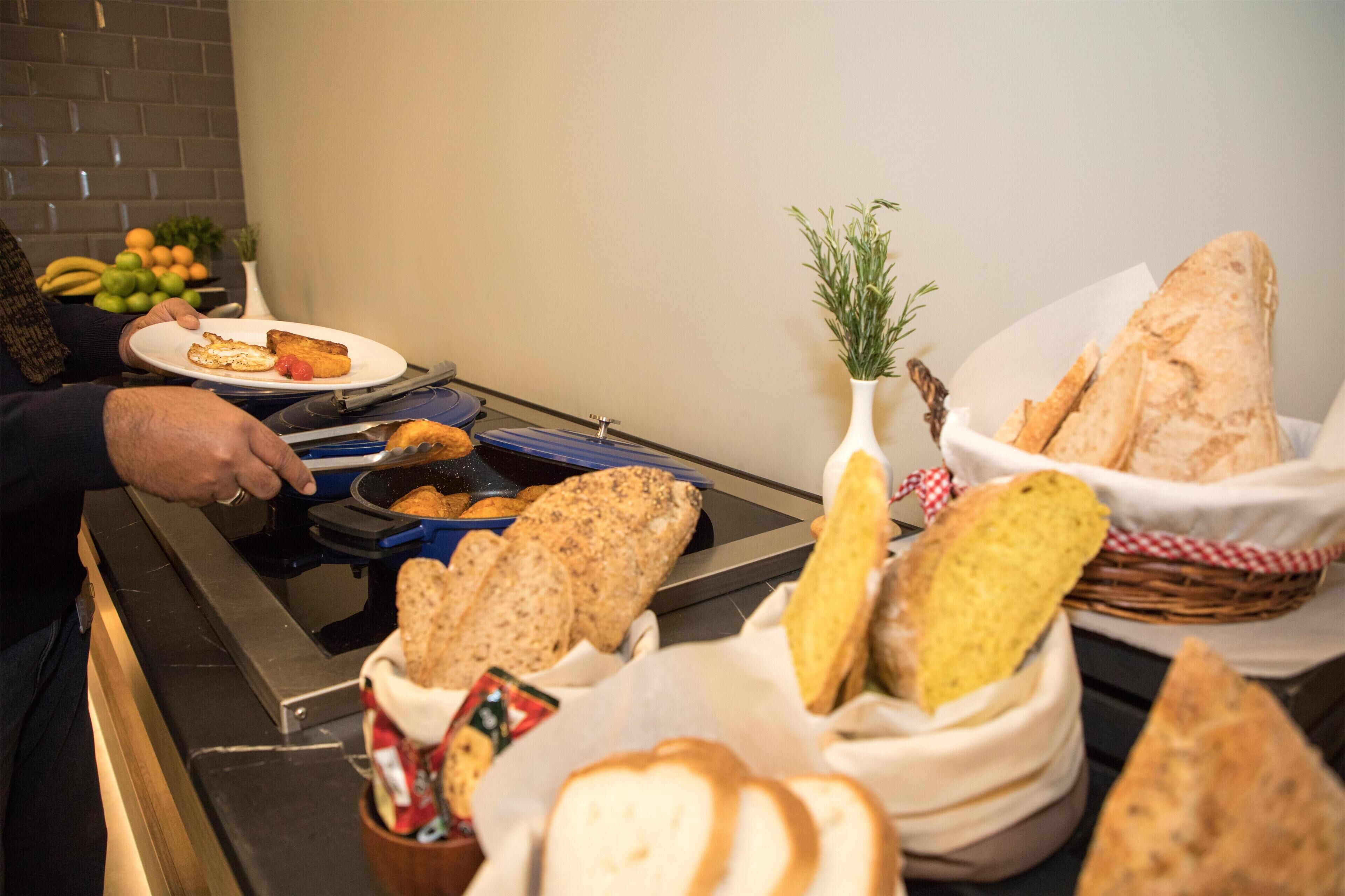Uma mesa de buffet com diversos alimentos, incluindo pão, um prato com carne e legumes, e uma cesta de mini pães.