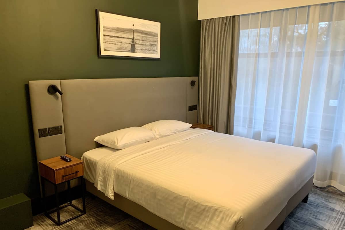Une chambre d’hôtel avec un grand lit, une table de chevet avec une télécommande, un tableau accroché au mur et une fenêtre avec des rideaux.