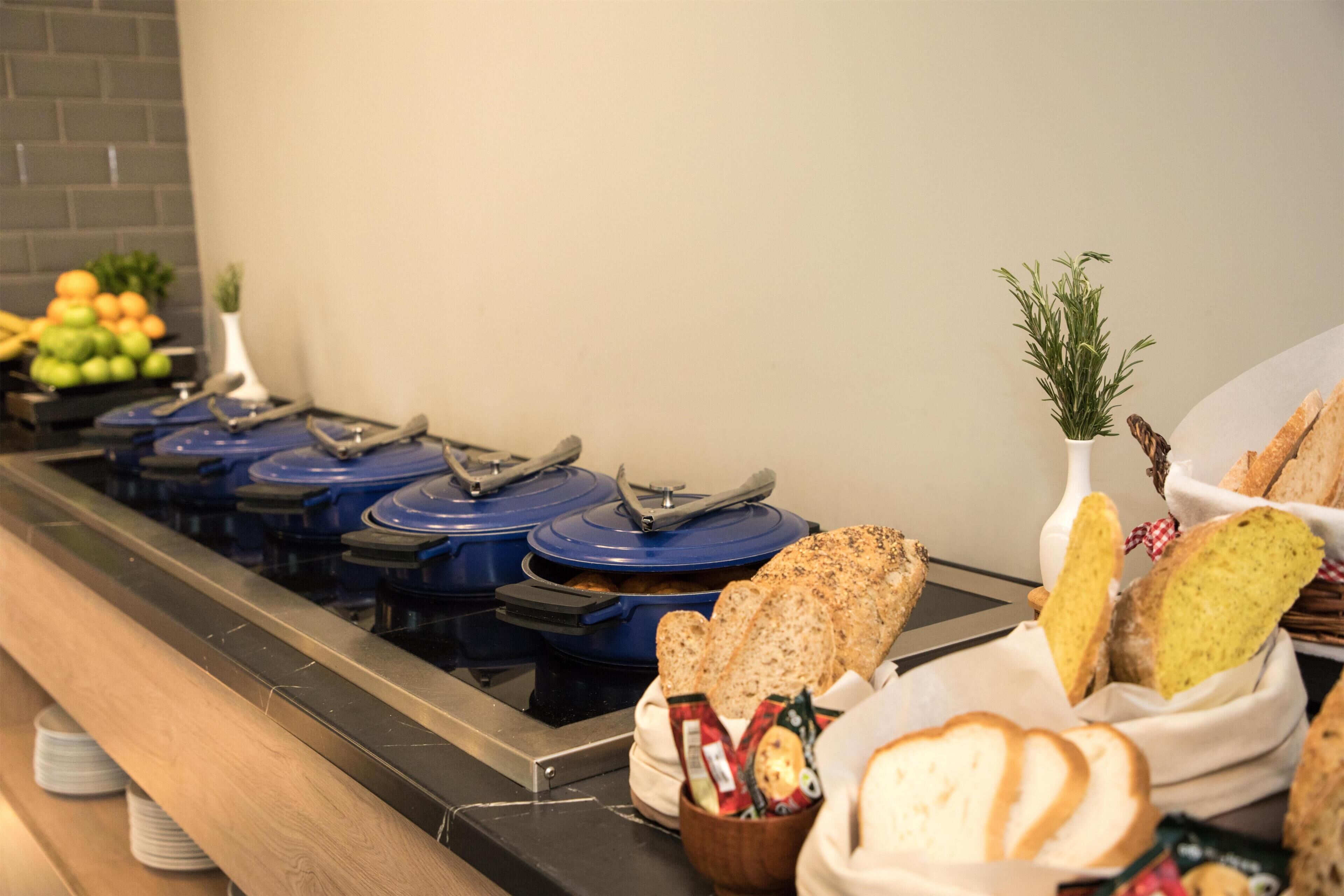 Disposição de buffet com diversas estações de comida, incluindo uma mesa de frutas e cestas de pão.