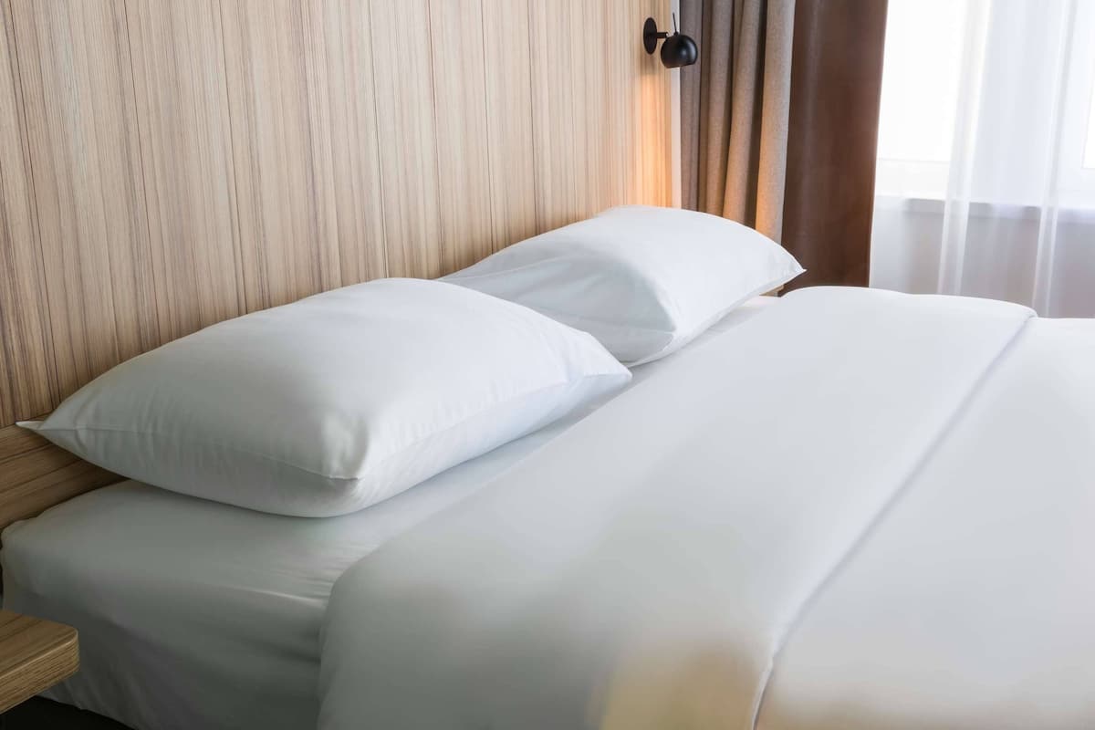 Un lit bien fait, avec du linge de lit blanc et des oreillers, une tête de lit en bois et une lampe noire fixée au mur.
