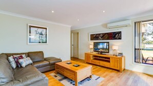 A living room with a brown leather sofa, a wooden coffee table, a TV on a wooden stand, and a large window with a view of the outdoors. - Peaceful Waterfront by Book Jervis Bay - wheelchair accessible - Stunning Location for Family Holiday (Sanctuary Point)