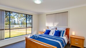 A bedroom with a large window, a bed with blue bedding, two bedside tables, and a view of a fenced outdoor area. - Peaceful Waterfront by Book Jervis Bay - wheelchair accessible - Stunning Location for Family Holiday (Sanctuary Point)