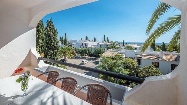 A balcony with wicker chairs, a table with a vase of flowers, and a view of a residential area with houses and trees. - Carolina Sea Apartment By Dreamplaces (Albufeira)