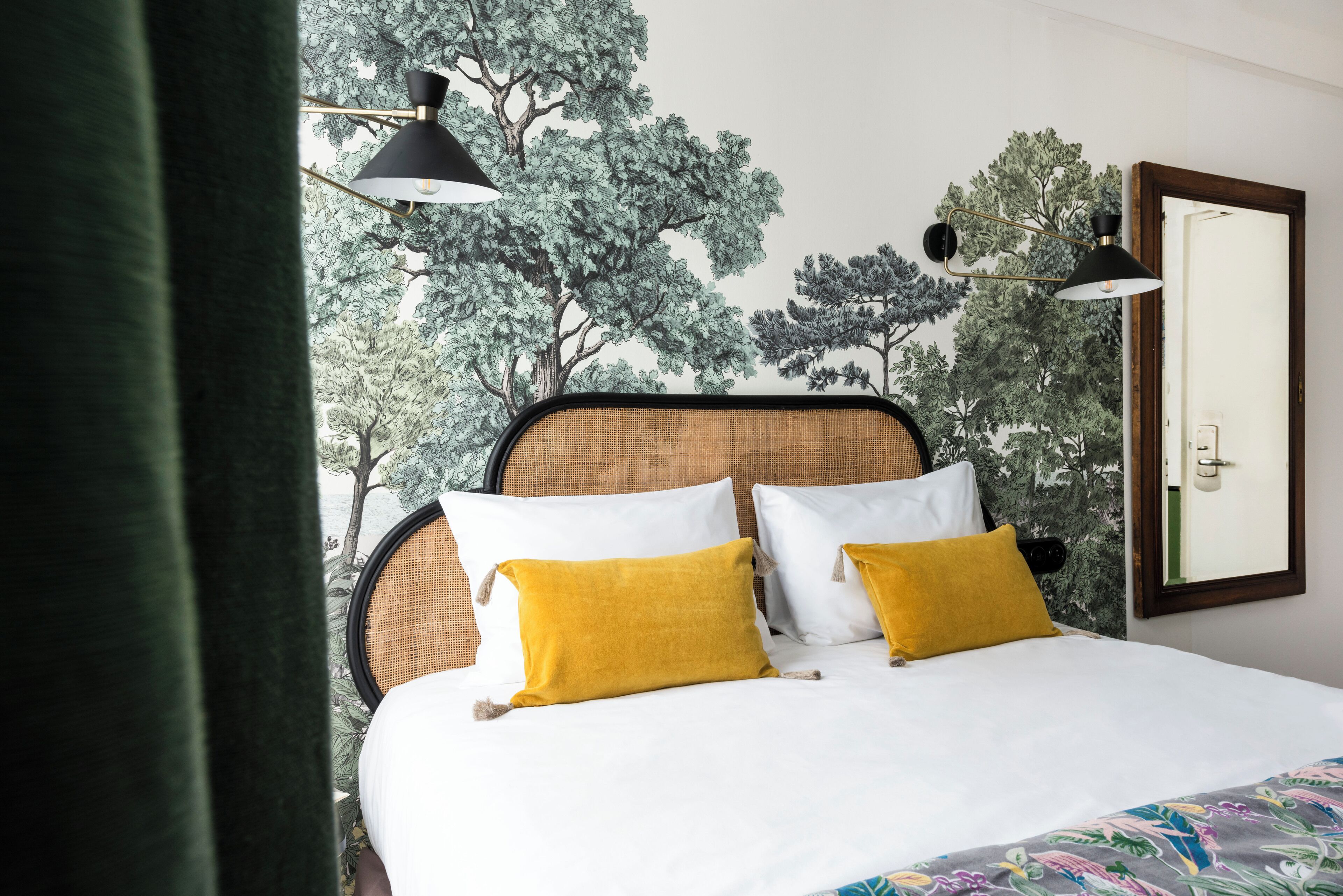 A bed with white and mustard pillows, a headboard with a woven pattern, and a wall mural of trees.