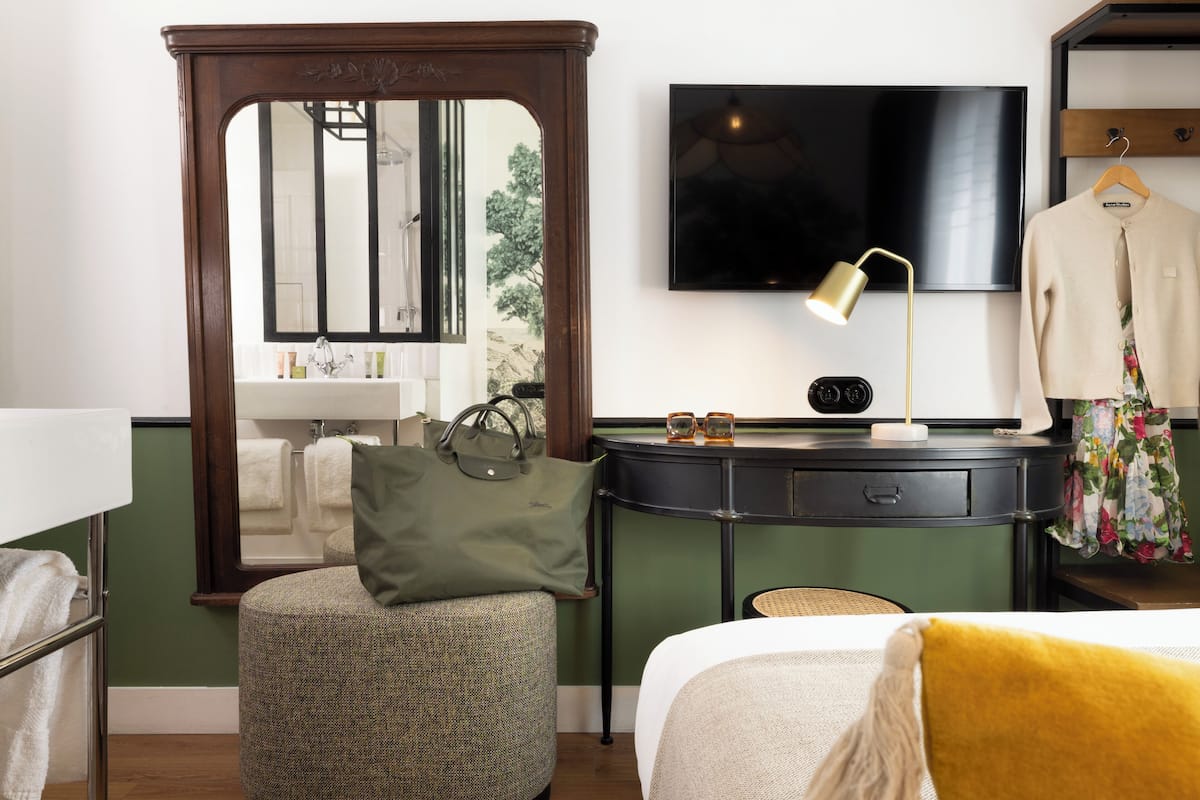 Habitación de hotel con cama con dosel, mesita de noche con lámpara, espejo, televisor montado en la pared y ventana con vistas a árboles.
