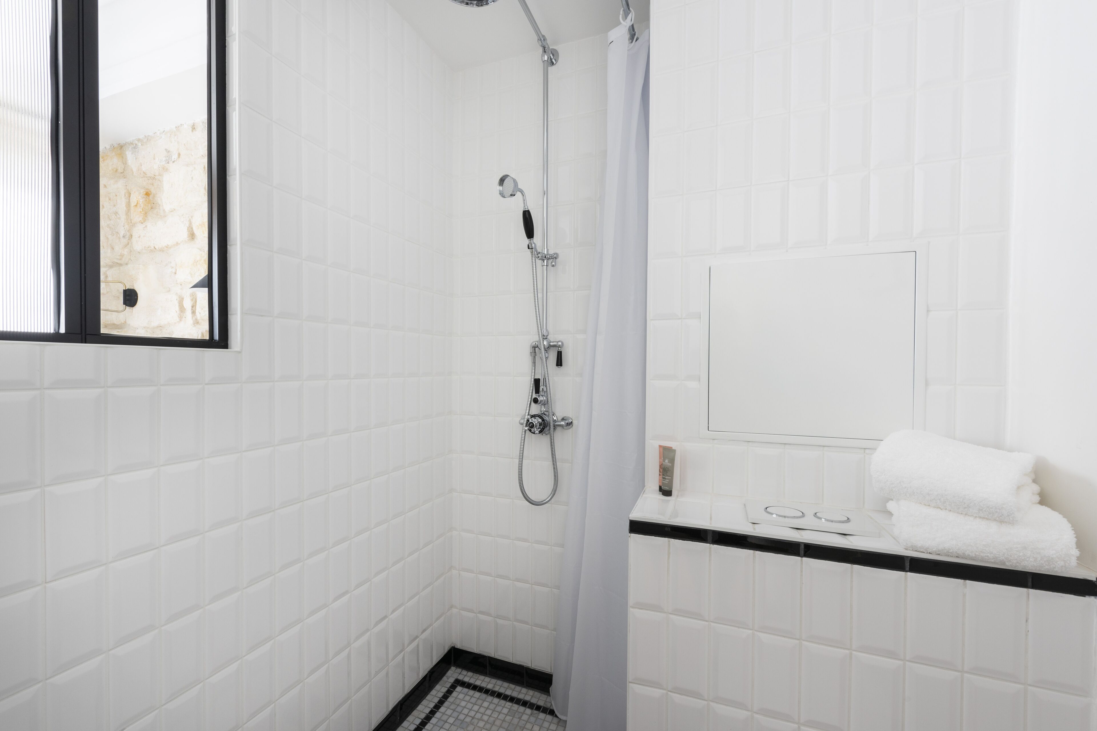 A small, white-tiled bathroom with a shower, a window, and a towel rack.