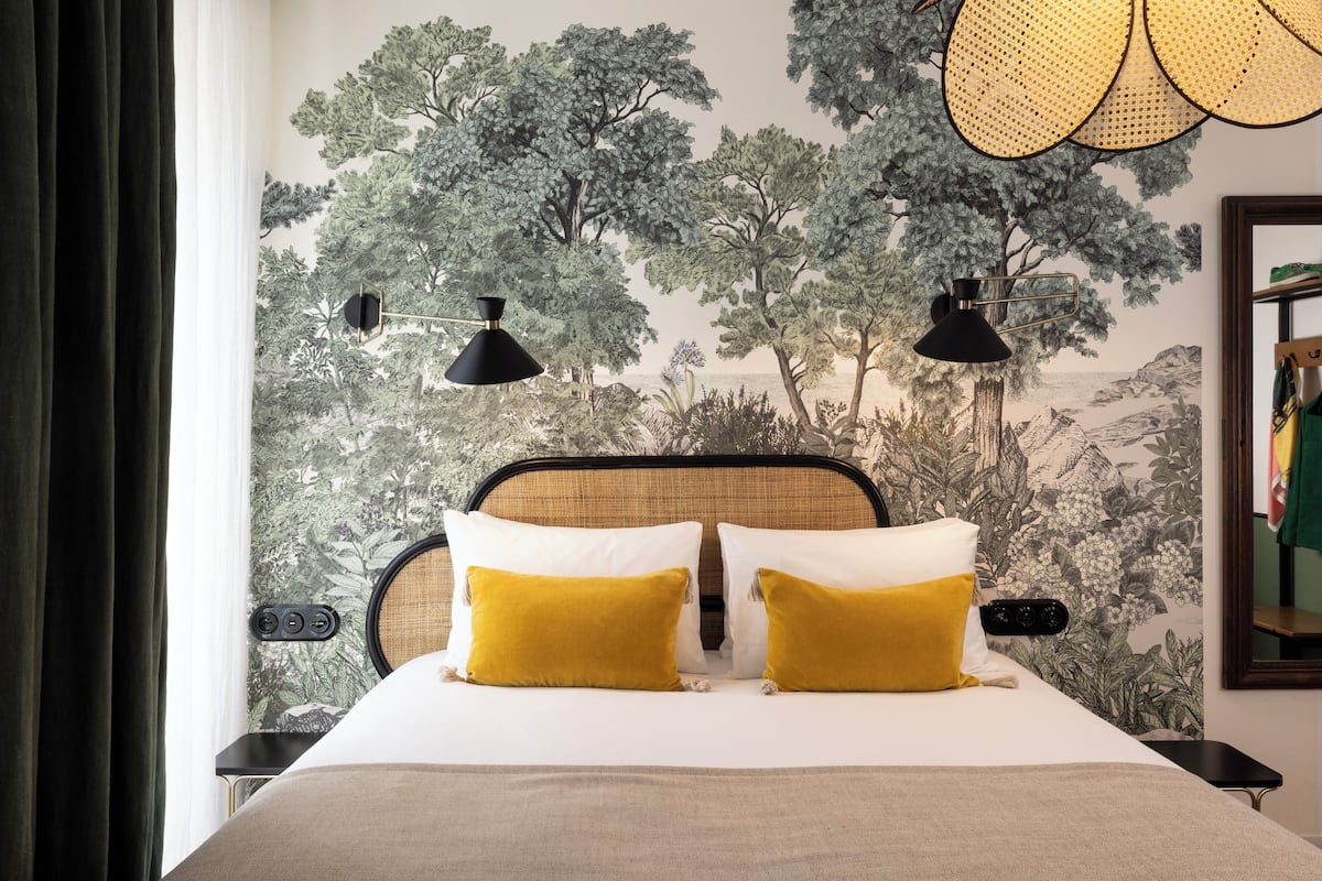 Una cama bien hecha con dos almohadas amarillas, un cabecero negro y un mural de árboles en la pared.