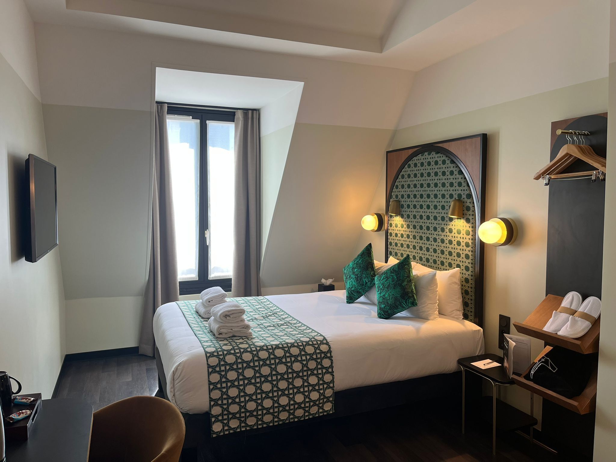 Een hotelkamer met een bed, een bureau, een stoel, een televisie, een raam met gordijnen en een wandplank met handdoeken.