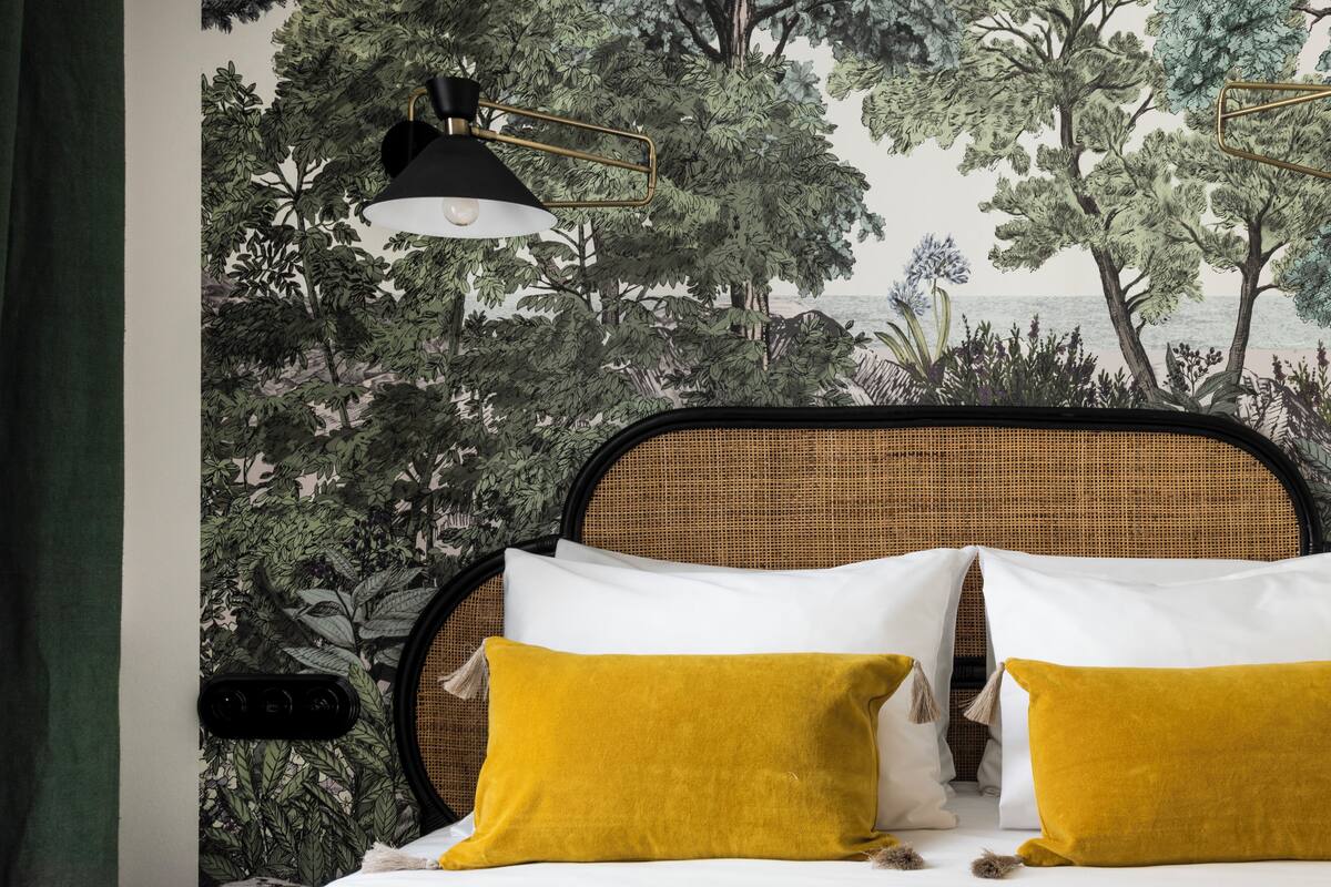 Una cama con almohadillas blancas y mostaza, una lámpara negra y un papel tapiz con motivos de naturaleza.
