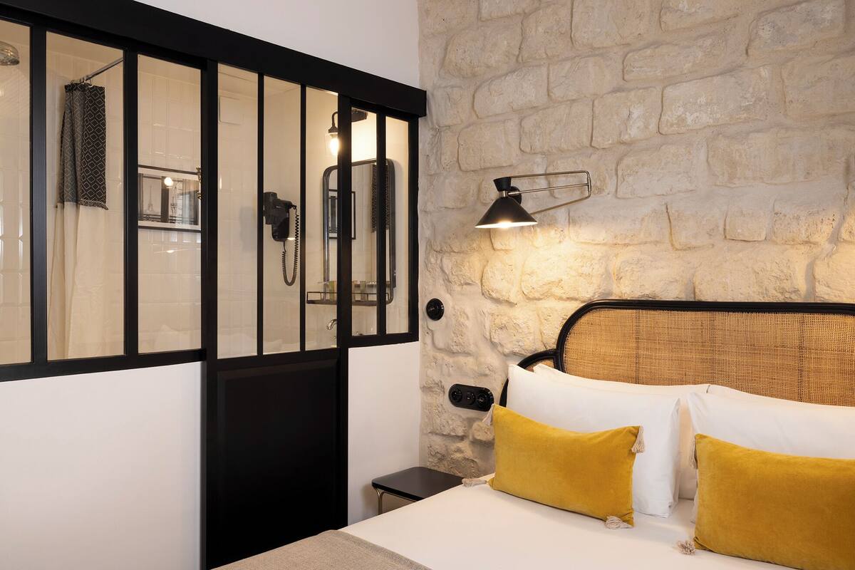 Un dormitorio con pared de piedra, cama con almohadas blancas y mostaza, mesita de noche, lámpara y zona de ducha con mampara de vidrio.
