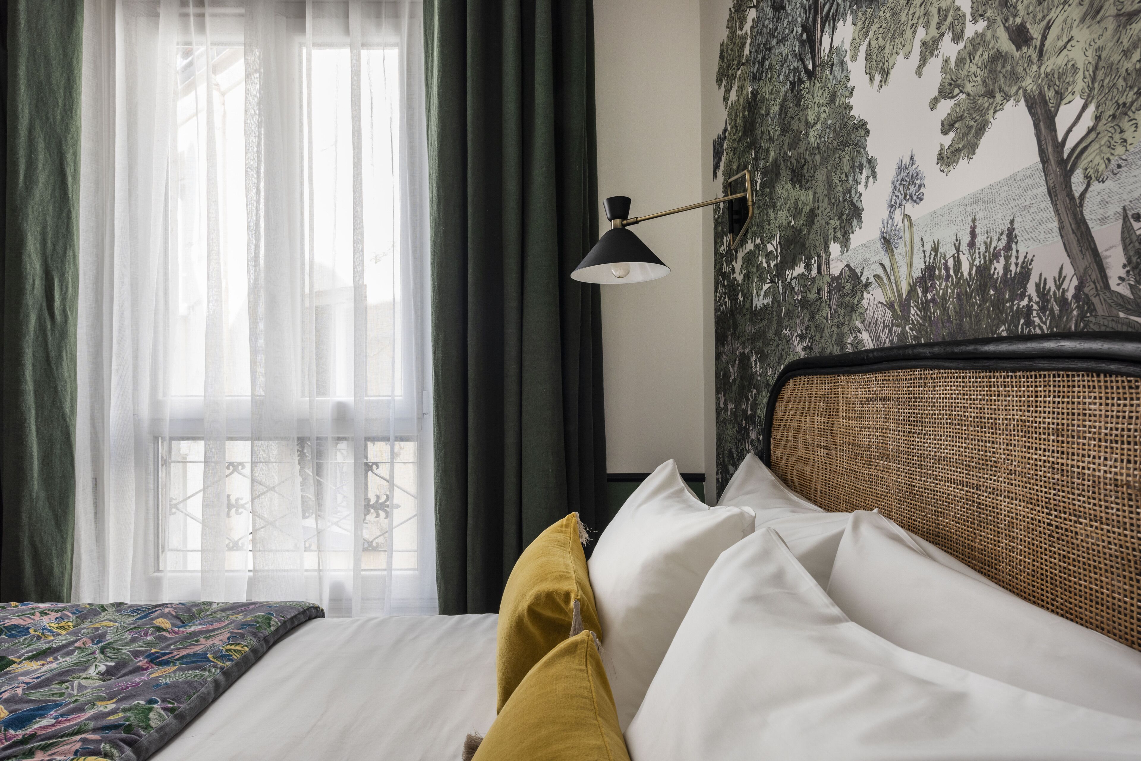 Un dormitorio con cama, una ventana con cortinas traslúcidas, una lámpara de pared y un mural con motivos de árboles.