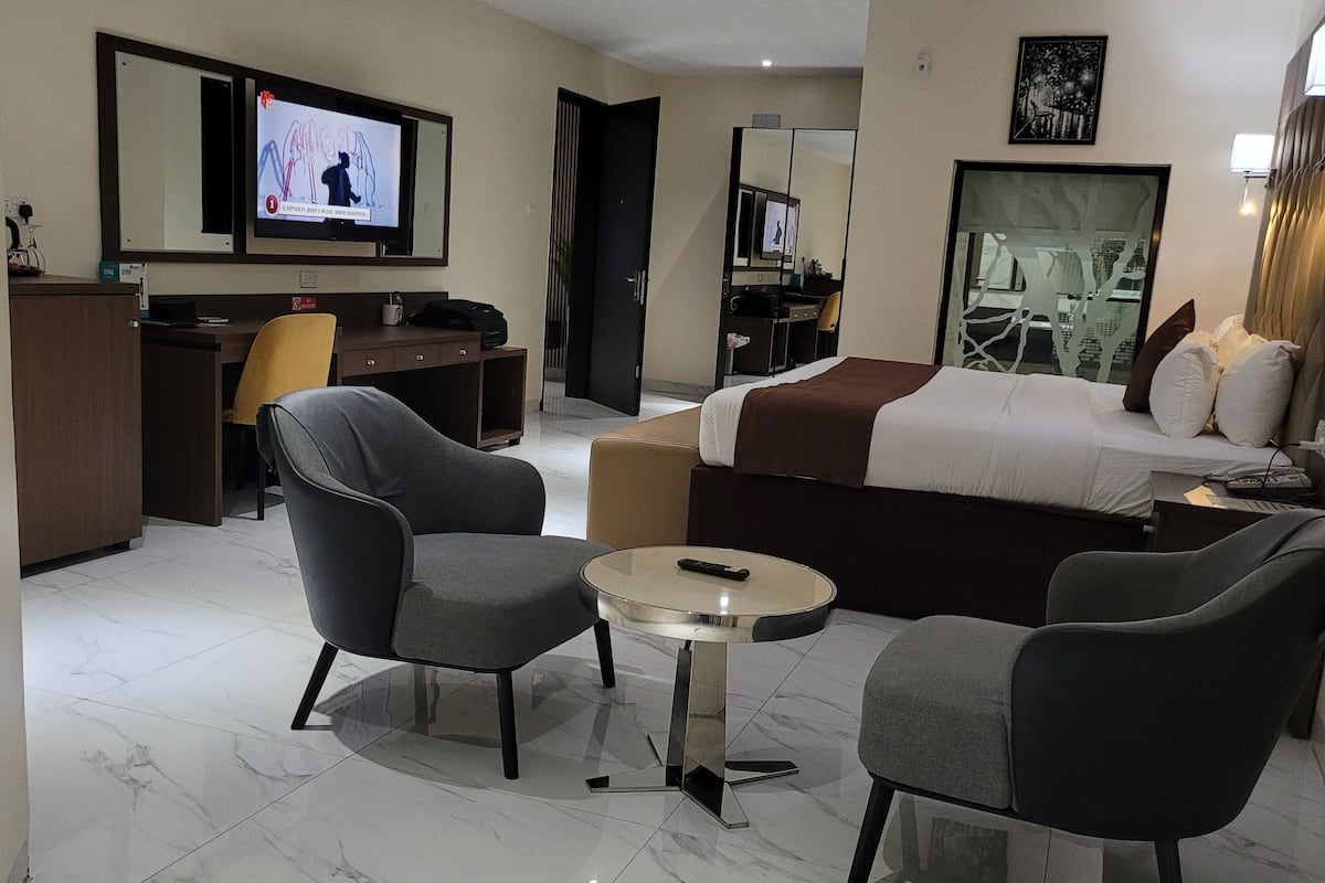 Une chambre d’hôtel moderne comprenant un lit, deux fauteuils, une petite table ronde, un bureau, une télévision et un miroir.