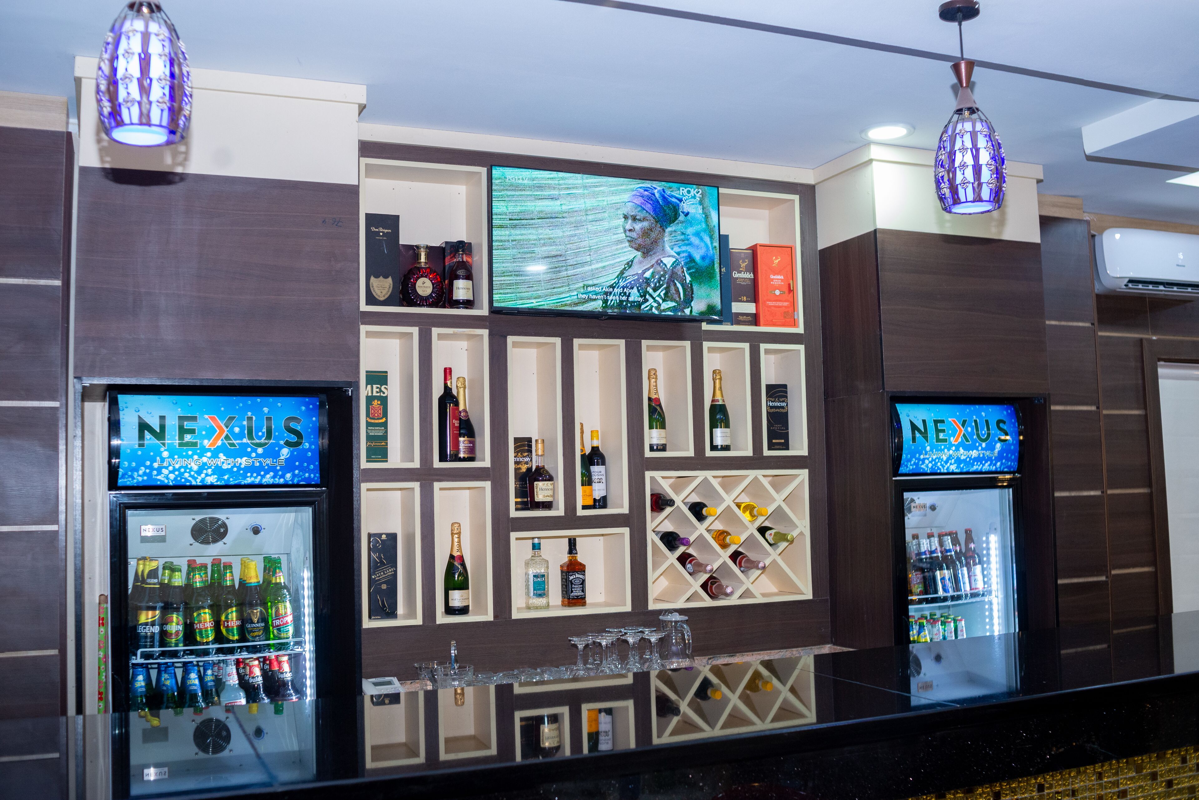 Un espace bar équipé de deux réfrigérateurs de la marque « NEXUS », remplis de boissons, d’un écran de télévision diffusant un programme et d’étagères contenant diverses bouteilles et verres.