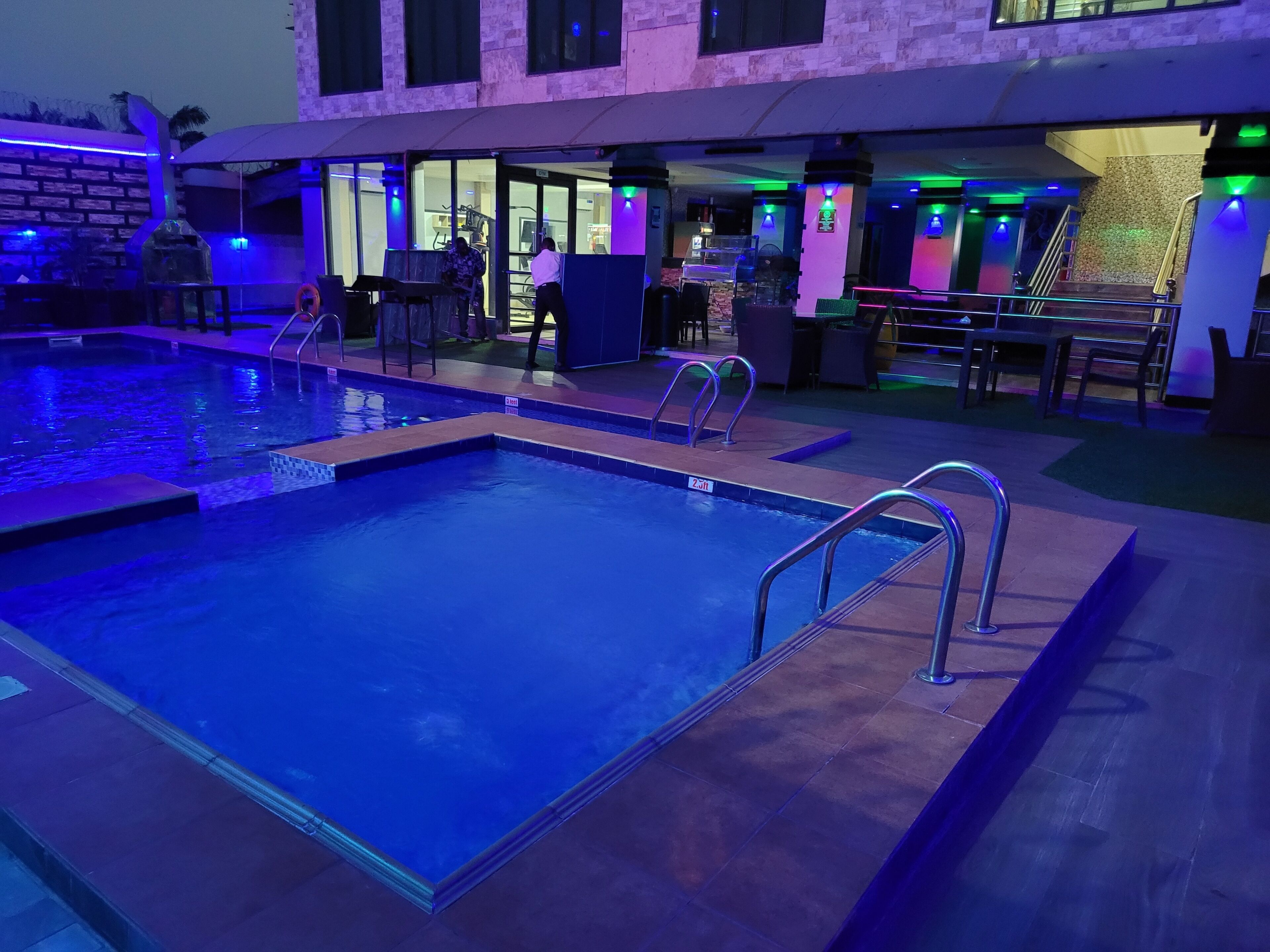 A swimming pool with a 2-jlt depth marker, surrounded by a lounge area with tables and chairs, and a building with glass doors and windows in the background.