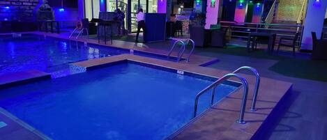A swimming pool with a 2-jlt depth marker, surrounded by a lounge area with tables and chairs, and a building with glass doors and windows in the background.