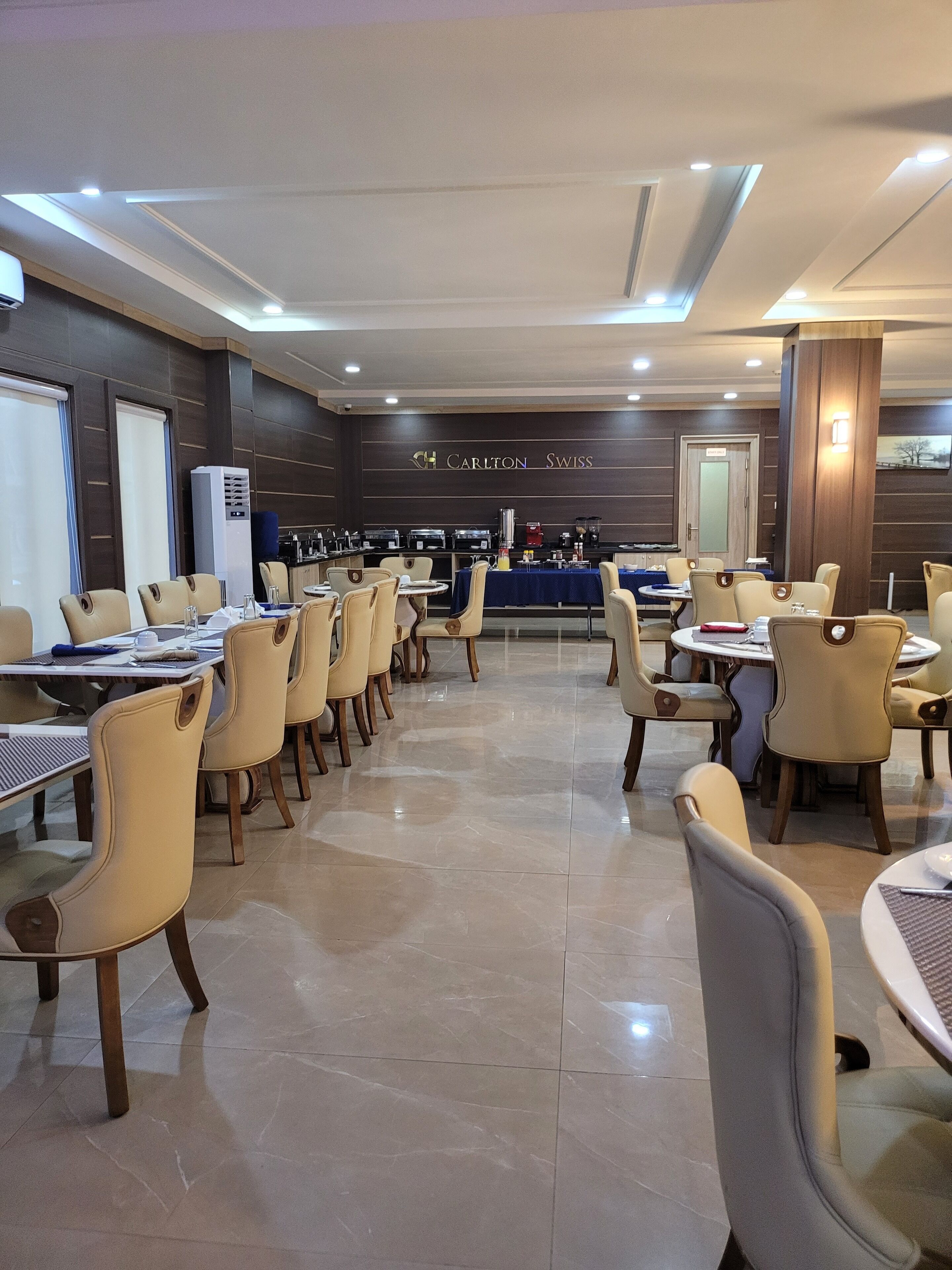 A hotel dining area with multiple tables and chairs, a kitchen area in the background, and a sign that reads "Carlton Swiss".