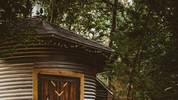 A rustic cabin with a distinctive star-shaped door, surrounded by a stone pathway and a forested area. - The Green Egg - Vintage RV Glamping (Terrell)