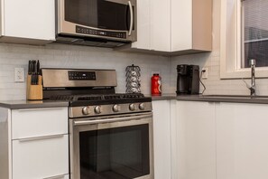 Uma cozinha com micro-ondas, fogão e forno de aço inoxidável. Há facas num bloco de madeira, um recipiente vermelho e uma cafeteira.