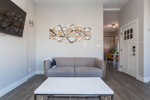 Uma sala de estar moderna com um sofá cinzento, uma mesa de centro branca, arte abstrata na parede e uma porta com uma placa.
