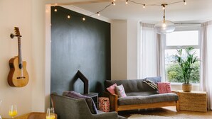 Un salon chaleureux avec un mur d’accent de couleur foncée, une guitare en bois, un canapé gris et une table à manger où sont disposés des aliments pour le petit-déjeuner.