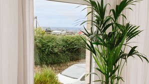 Une pièce dotée d’une grande fenêtre, d’une plante en pot, d’un canapé recouvert d’un plaid et d’une petite table sur laquelle repose un set de table en fibres tressées.