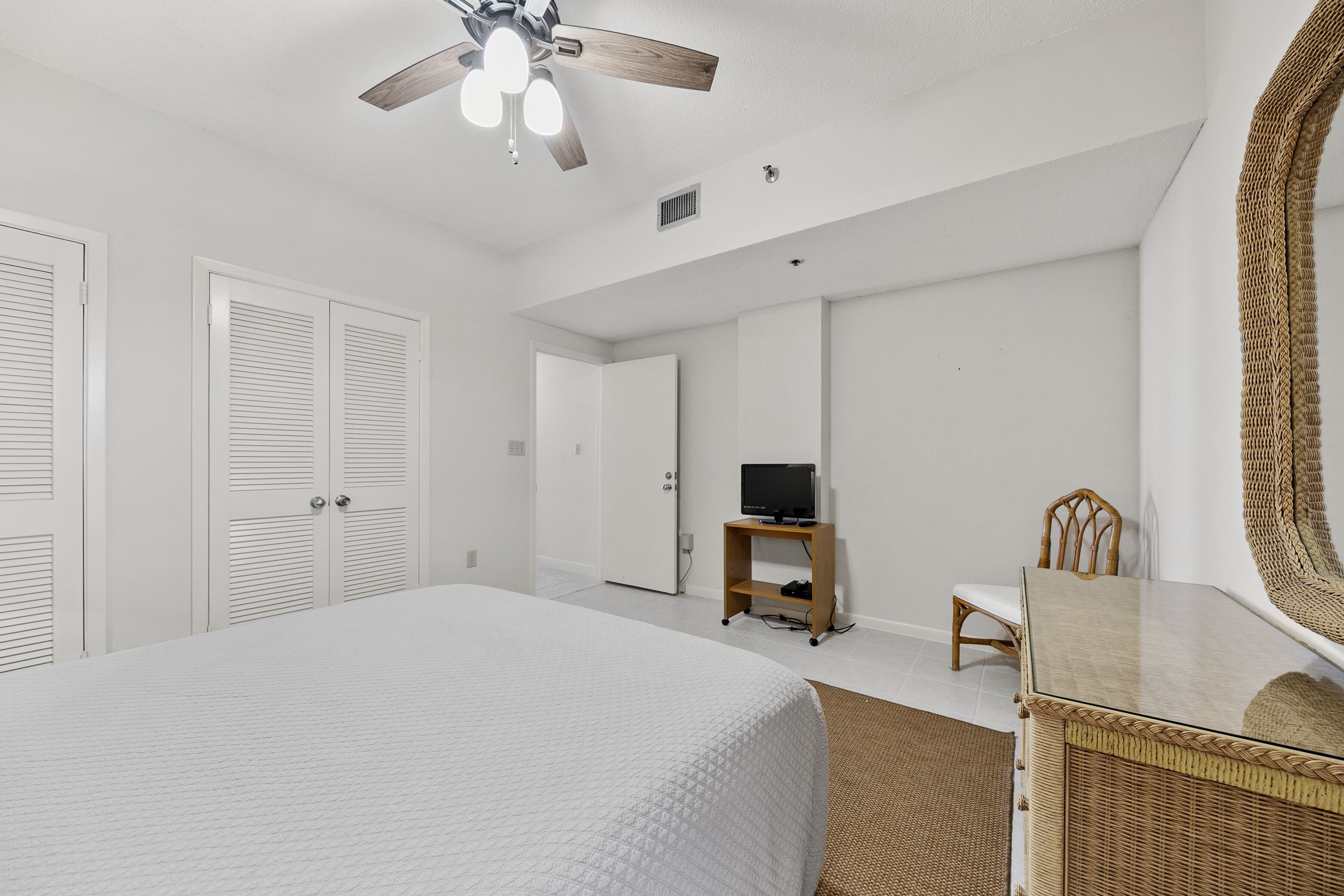 A bedroom with a large bed, a ceiling fan, a television on a stand, a wicker chair, and a large mirror.