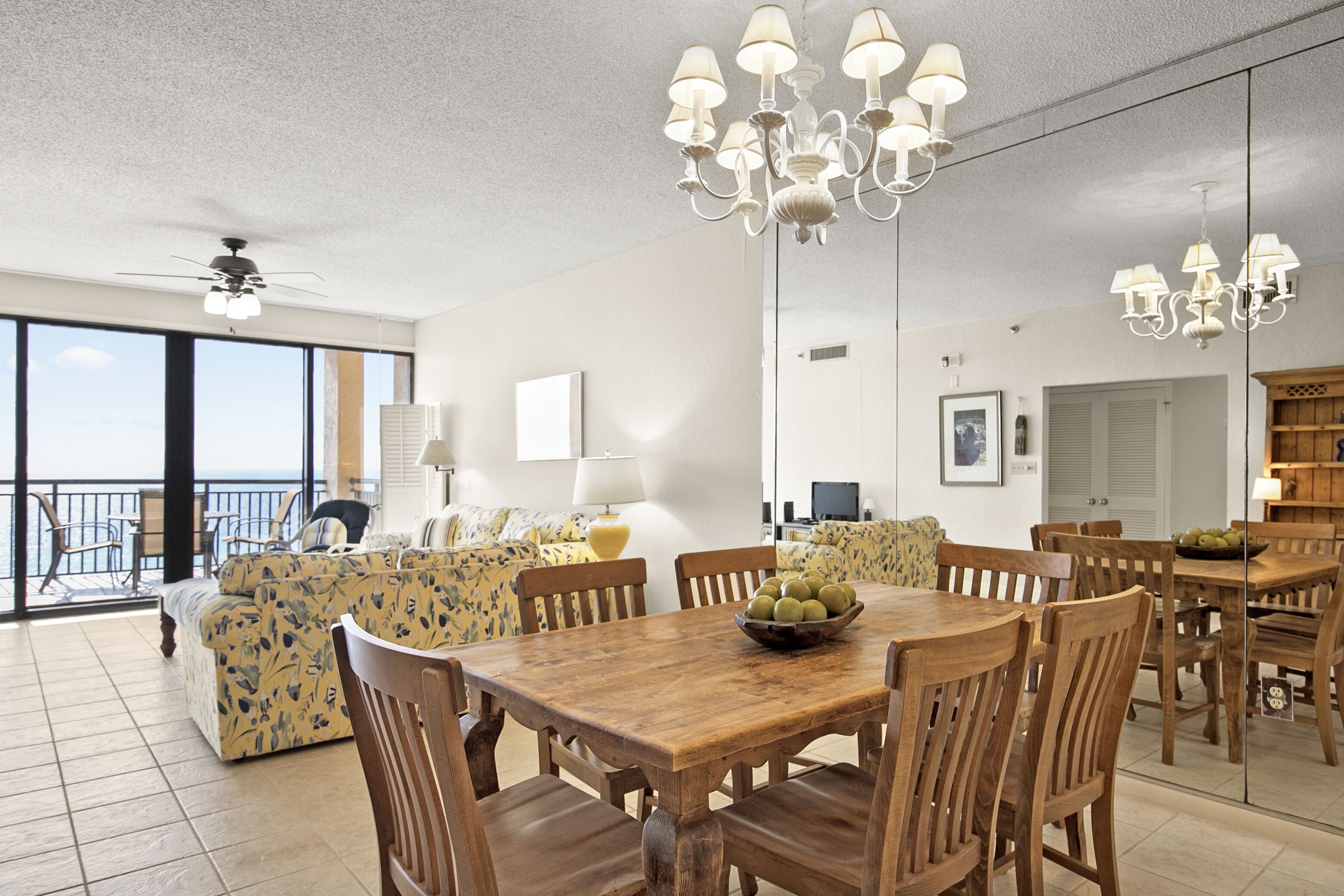 A spacious living room with a large dining table, wooden chairs, and a chandelier. There is a sofa with floral upholstery and a balcony with a view.