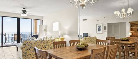 A spacious living room with a large dining table, wooden chairs, and a chandelier. There is a sofa with floral upholstery and a balcony with a view.