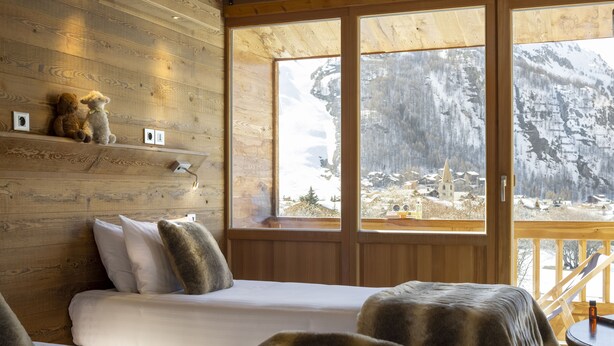 A cozy cabin room with a wooden interior, a bed, a chair, and a view of a snowy mountain landscape through large windows. - Hotel Ski Lodge (Val-d'Isere)