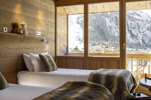 A cozy cabin room with a wooden interior, a bed, a chair, and a view of a snowy mountain landscape through large windows. - Hotel Ski Lodge (Val-d'Isere)