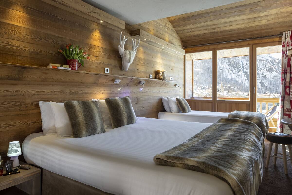 A hotel room with wooden walls, a large bed with white linens, a fur throw, a mounted deer head, and a window with a view of mountains.