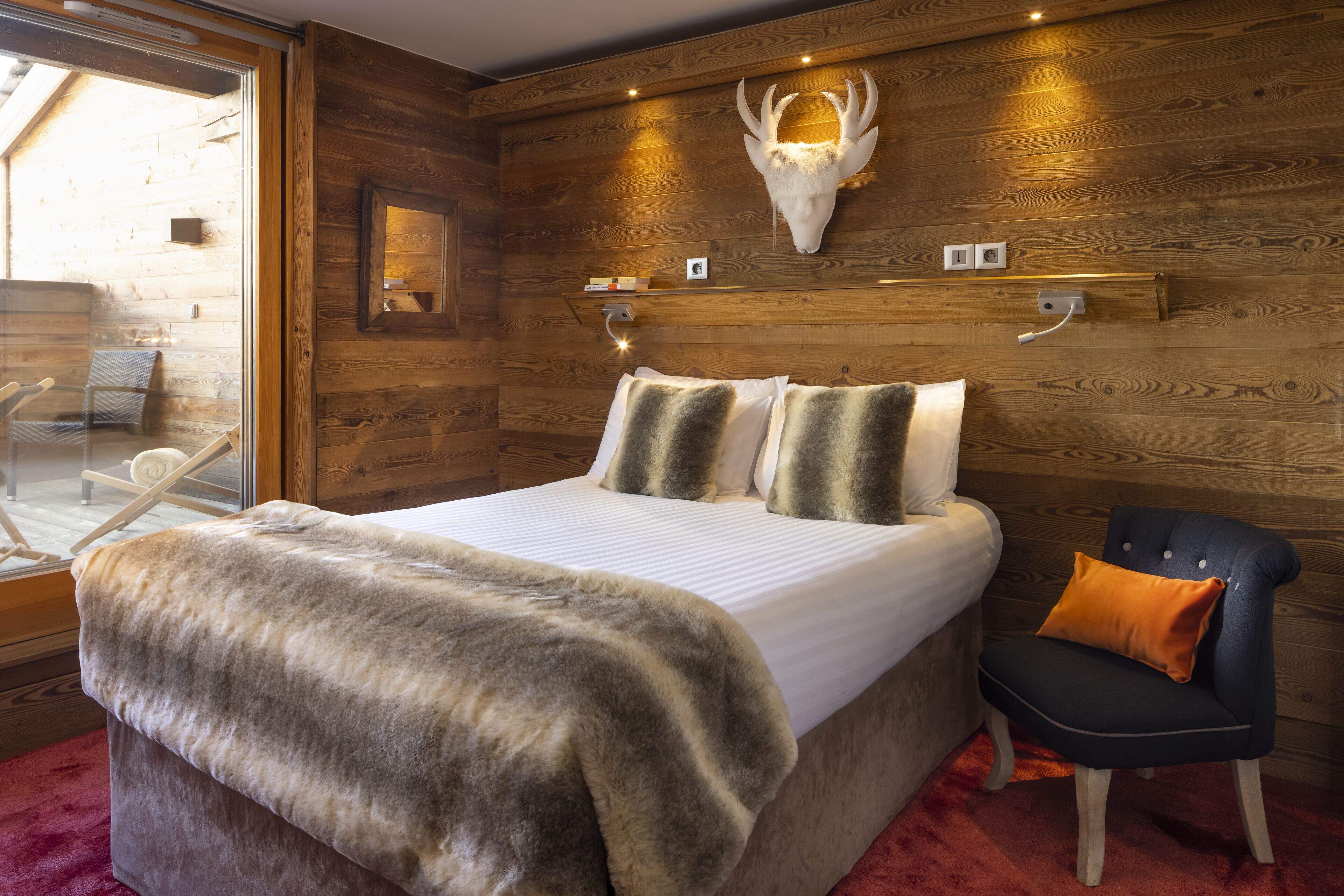 A wooden room with a bed, a chair, and a mounted animal skull on the wall.