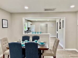 A dining area with a table set for four, a kitchen in the background, and a hallway leading to another room.