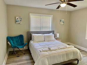 Ein Schlafzimmer mit einem Bett, Deckenventilator, Fenster mit Jalousien, einem Sessel und Kunstwerken an den Wänden.
