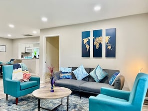 Ein modernes Wohnzimmer mit einem grauen Sofa, Türkis-farbenen Sesseln und einem Couchtisch. An den Wänden hängen drei Weltkarten.