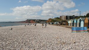 A pebbled beach with people sunbathing and walking, a row of colorful beach huts, and buildings in the background. - Mackerel Cottage (Budleigh Salterton)