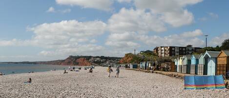 A pebbled beach with people sunbathing and walking, a row of colorful beach huts, and buildings in the background.