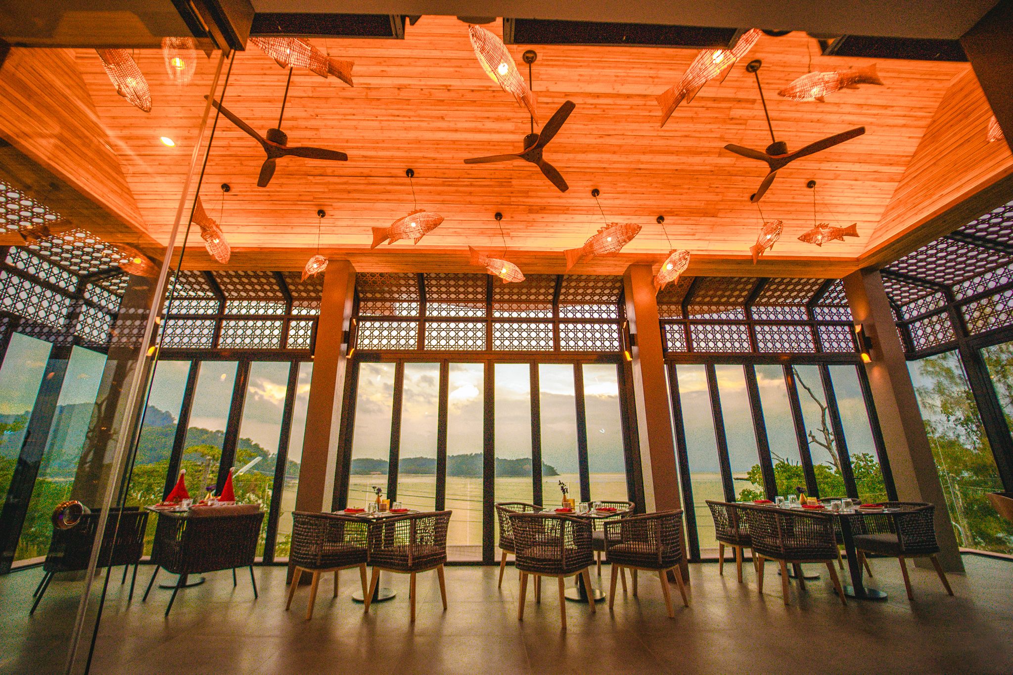 A dining area with tables and chairs, large windows with a view, and ceiling fans.