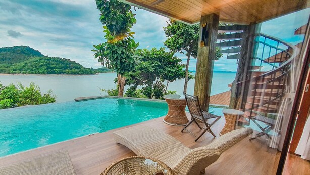 Un espace aménagé au bord de la piscine, avec des chaises longues et une balustrade en verre donnant sur une étendue d’eau et une végétation luxuriante.