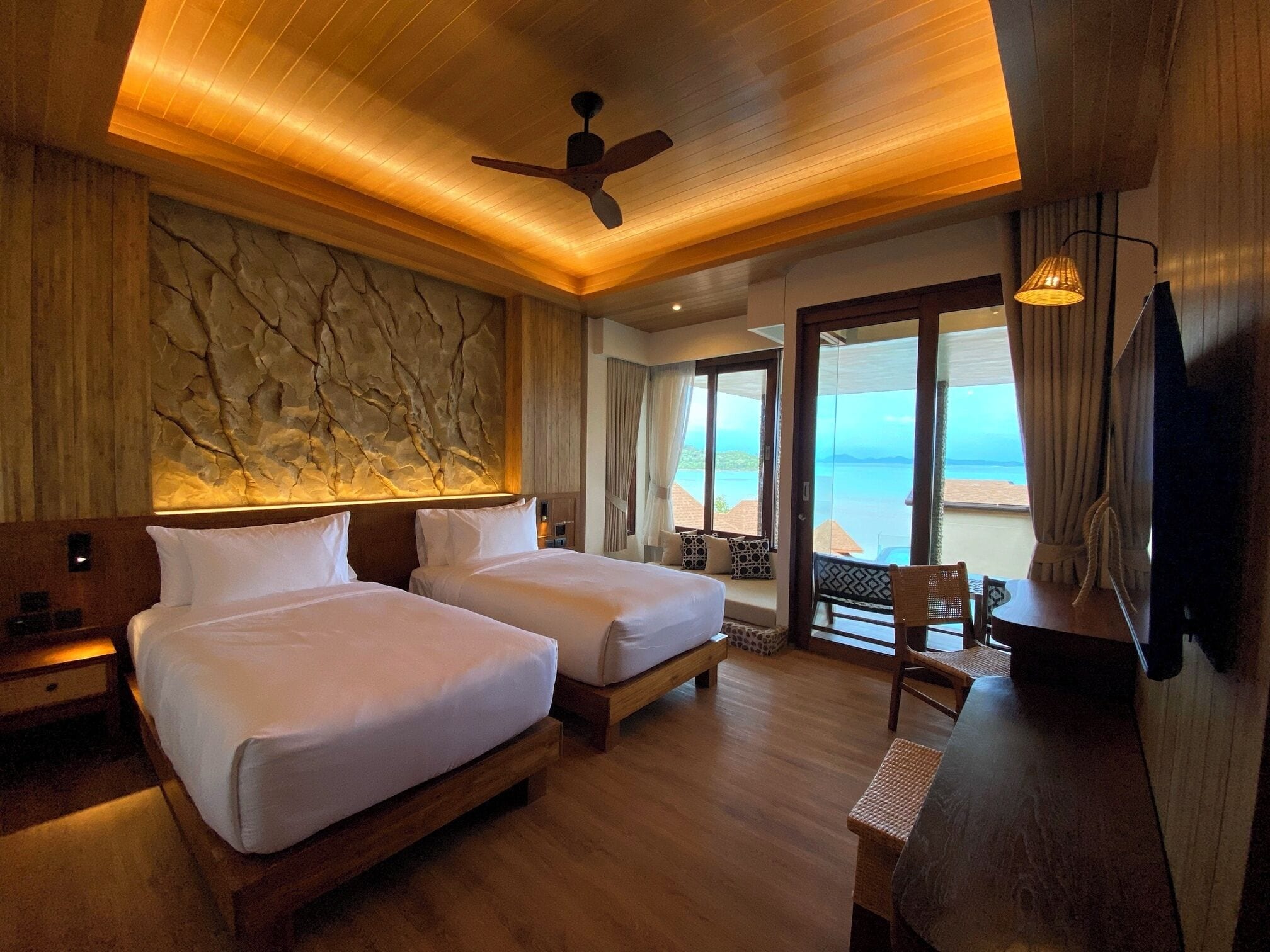A hotel room with two beds, a ceiling fan, a large window with a view of the beach, and a television.