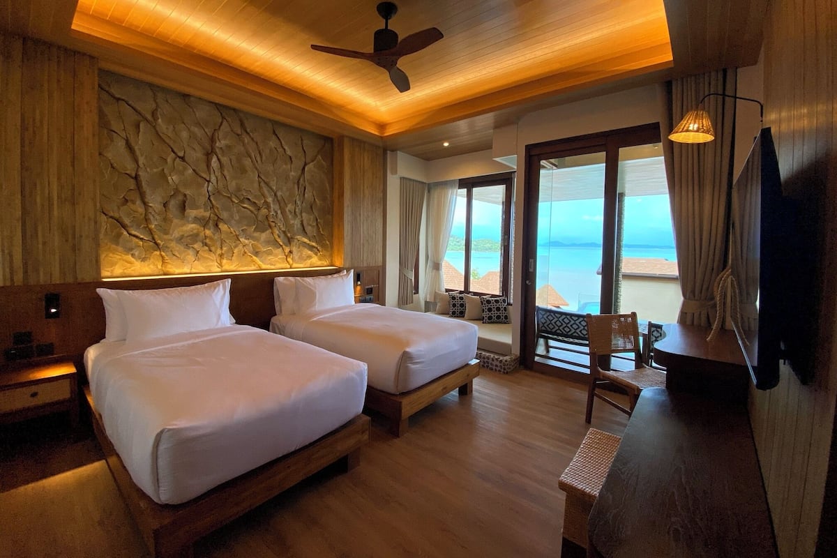 A hotel room with two beds, a ceiling fan, a large window with a view of the beach, and a television.