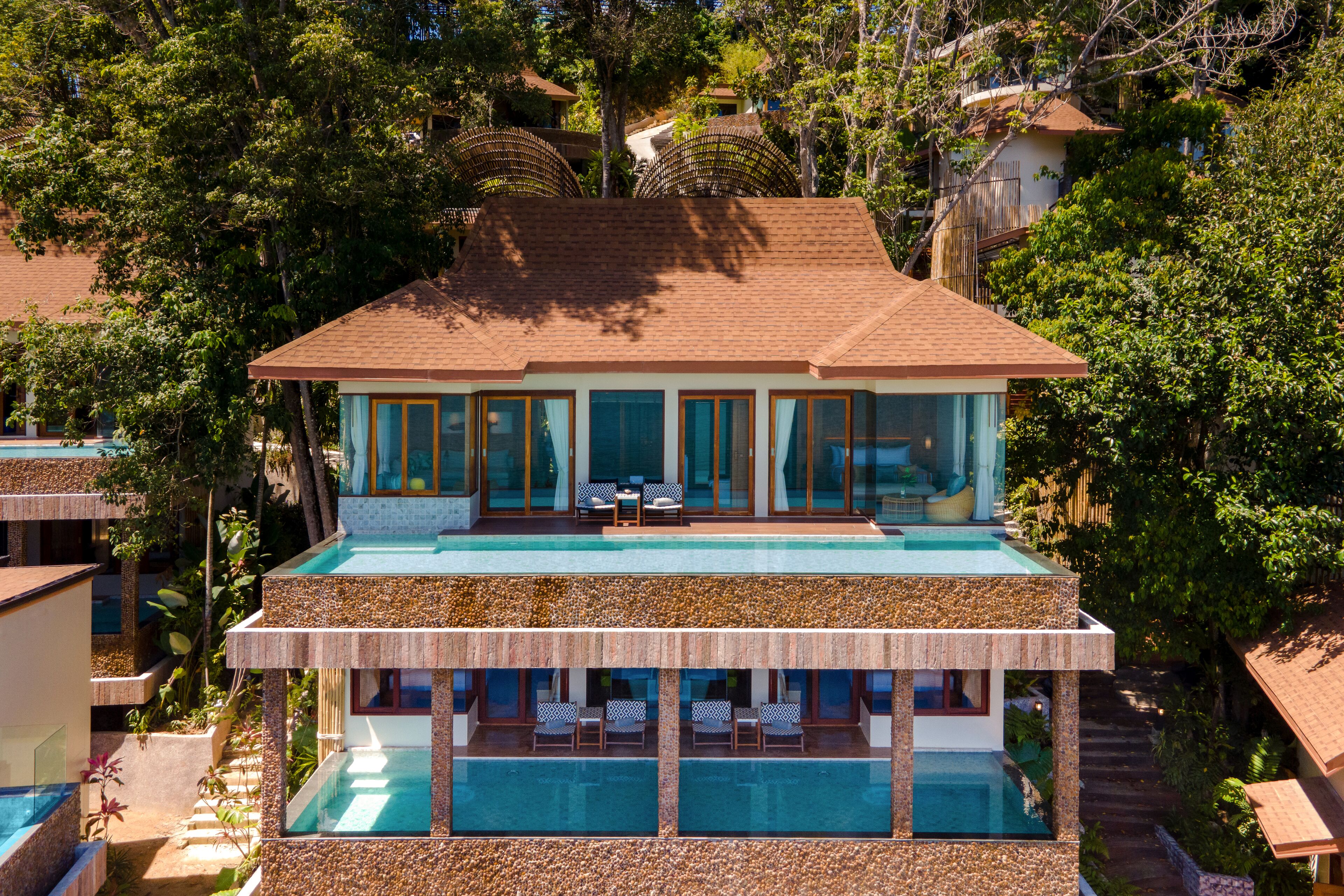 A modern two-story house with a large swimming pool on the ground floor and a balcony with outdoor seating.