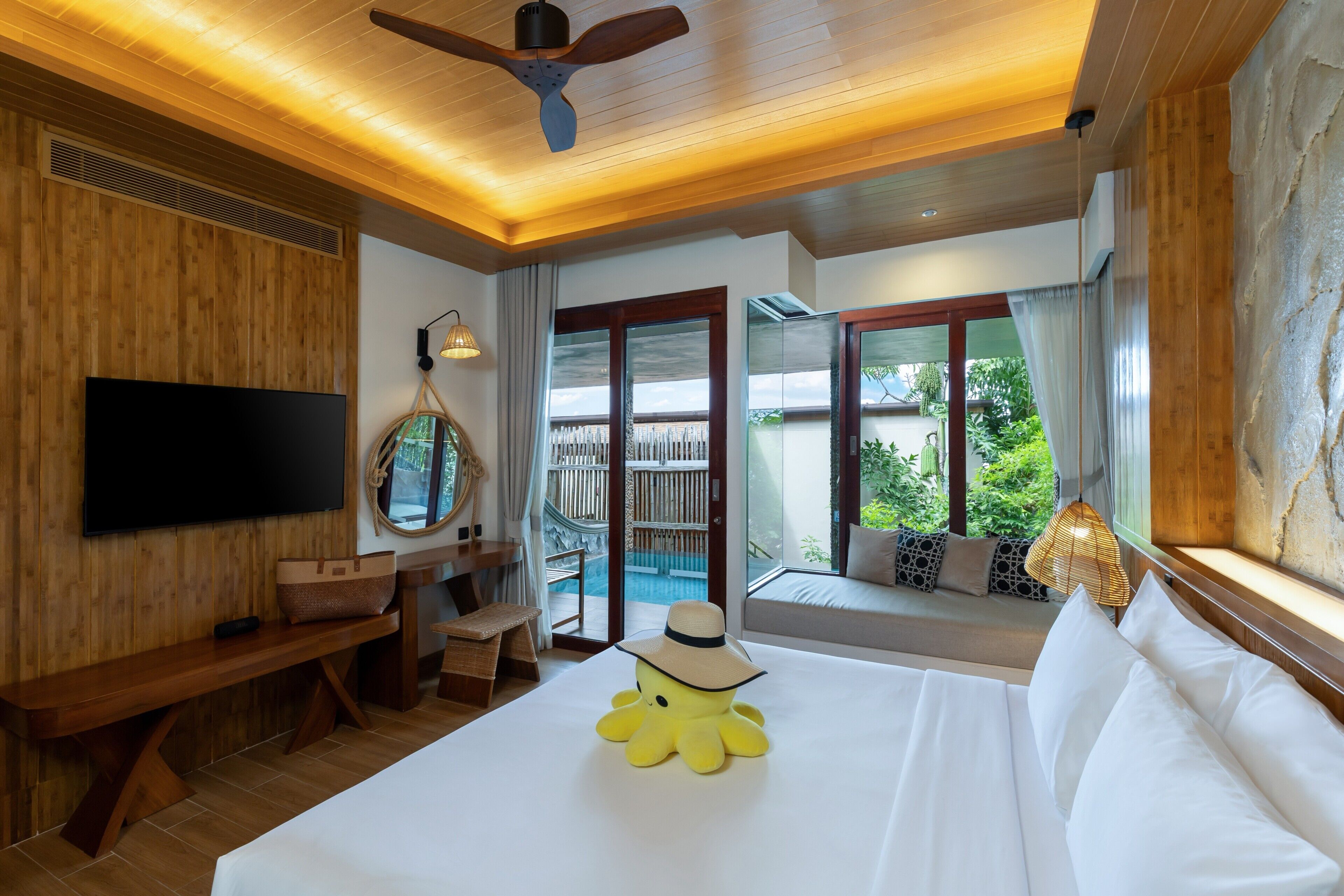 Habitación de hotel con una cama grande, televisión, ventilador de techo, ventana con vista al exterior y una pequeña zona de estar con vista a la piscina.