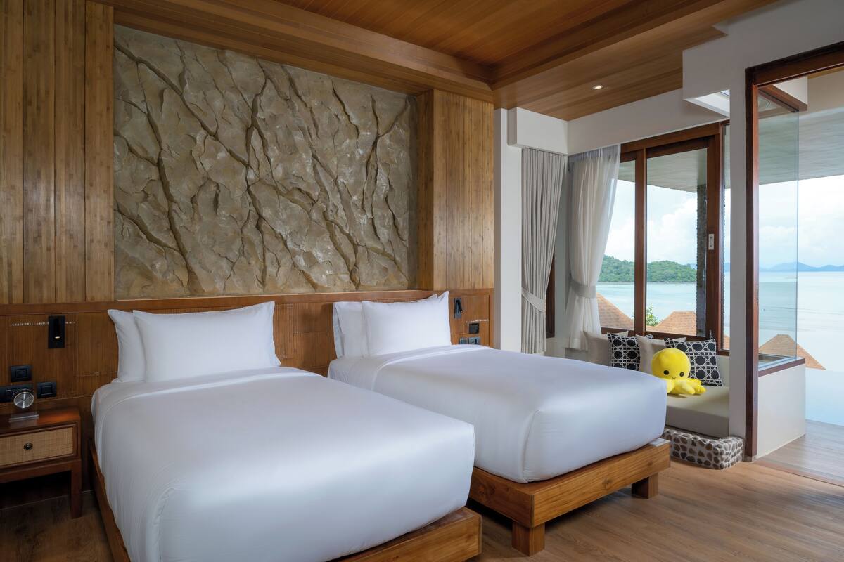 A hotel room with two beds, a stone wall, wooden ceiling, and a view of the sea.