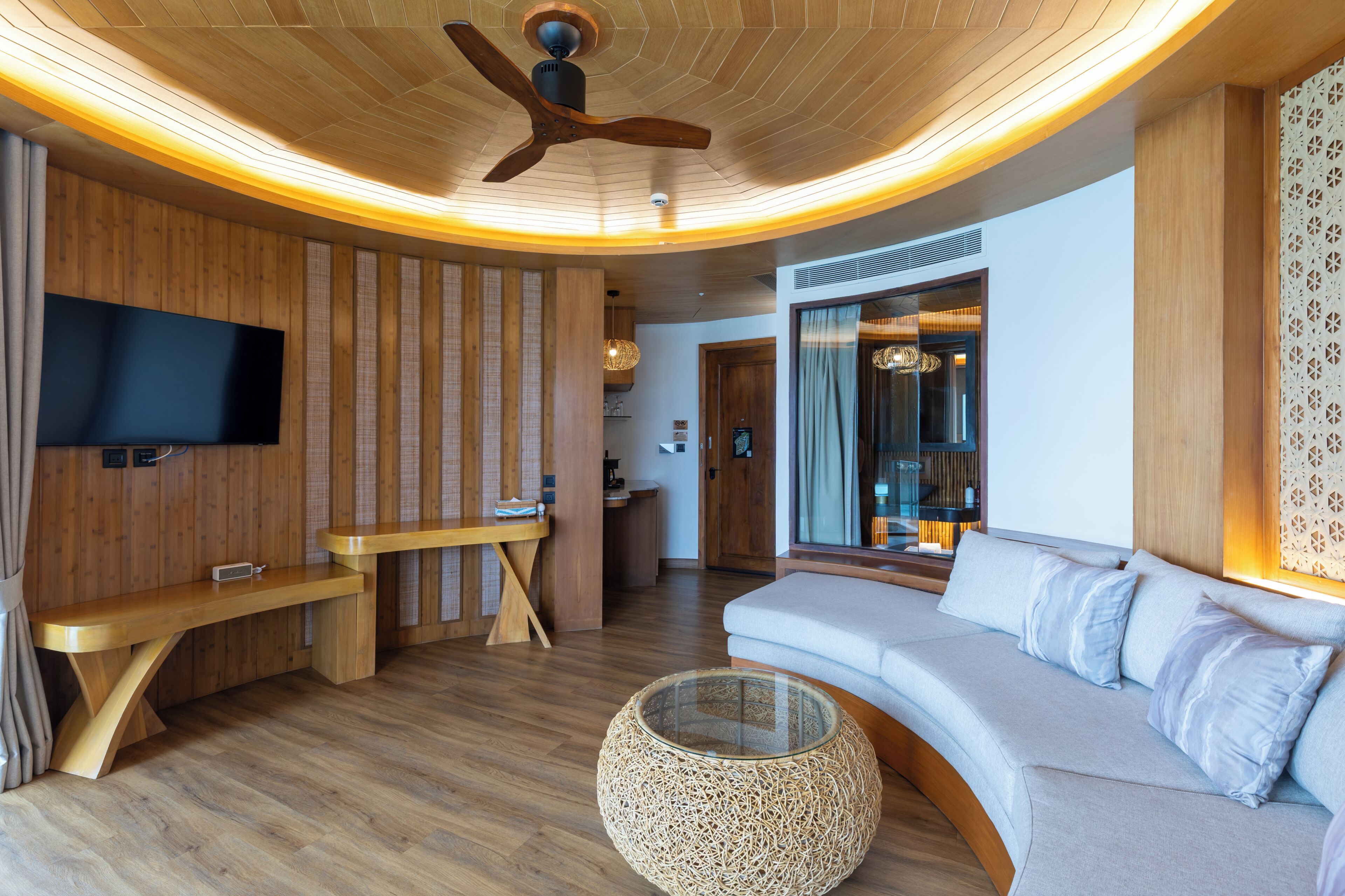 A modern living room with a curved wooden ceiling, a flat-screen TV mounted on the wall, a wooden bench, a round woven coffee table, and a light-colored sofa with cushions.