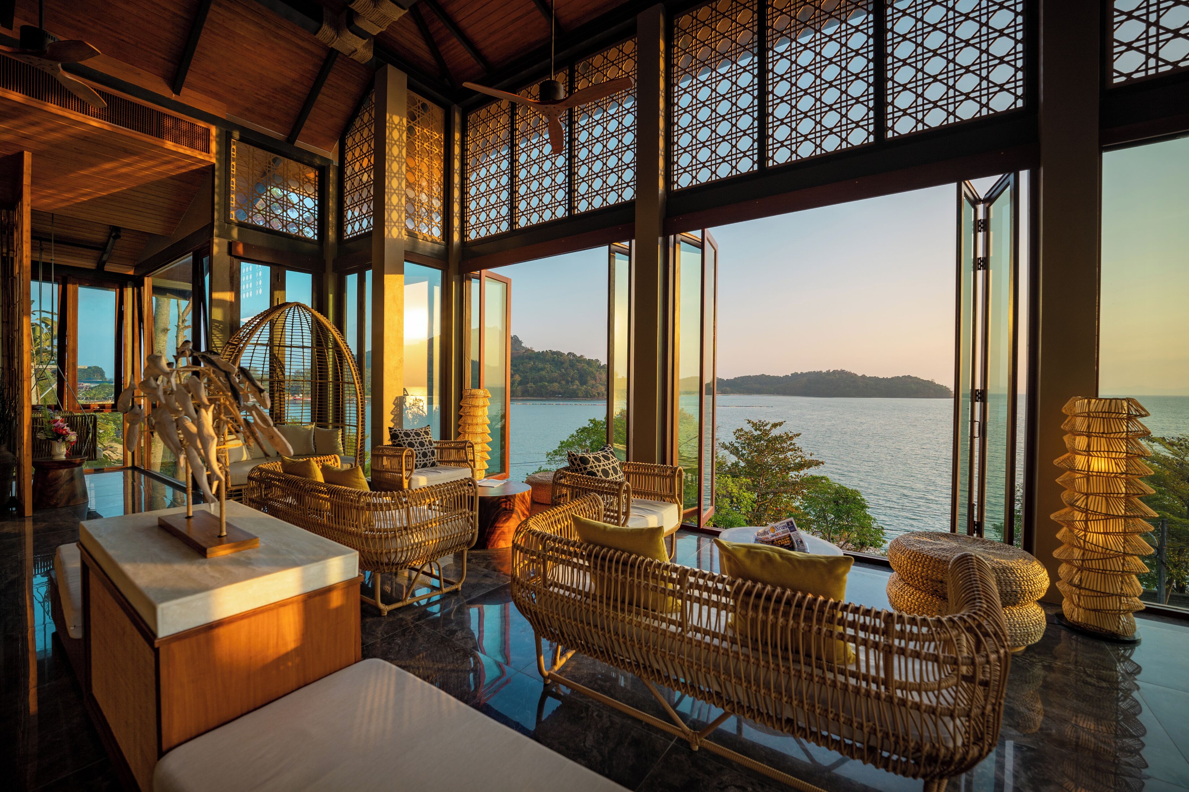 A spacious room with a large window overlooking a body of water, furnished with wicker furniture and a glass coffee table.