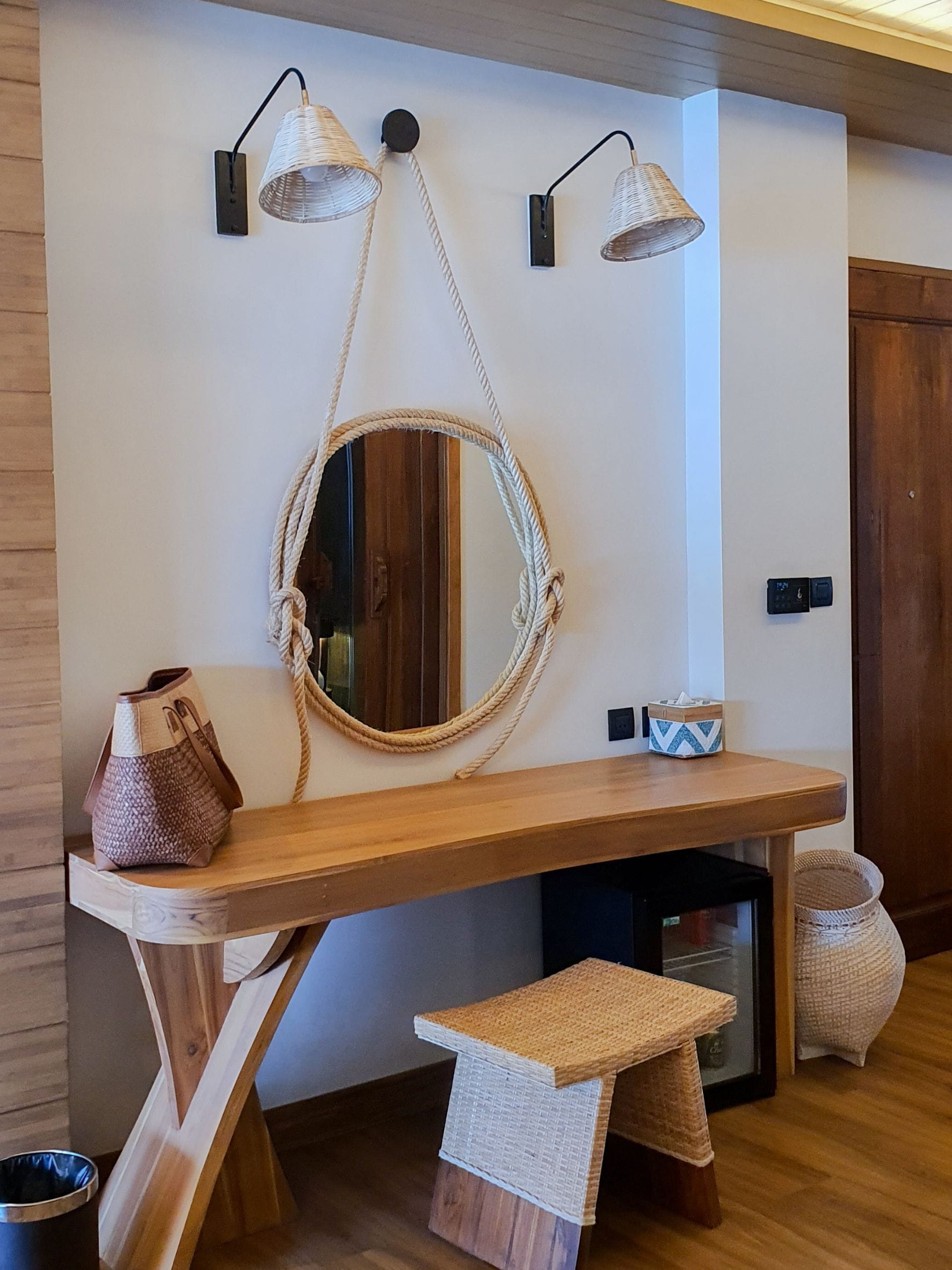 A wooden console table with a mirror, two wall-mounted lamps, a woven basket, and a small stool.