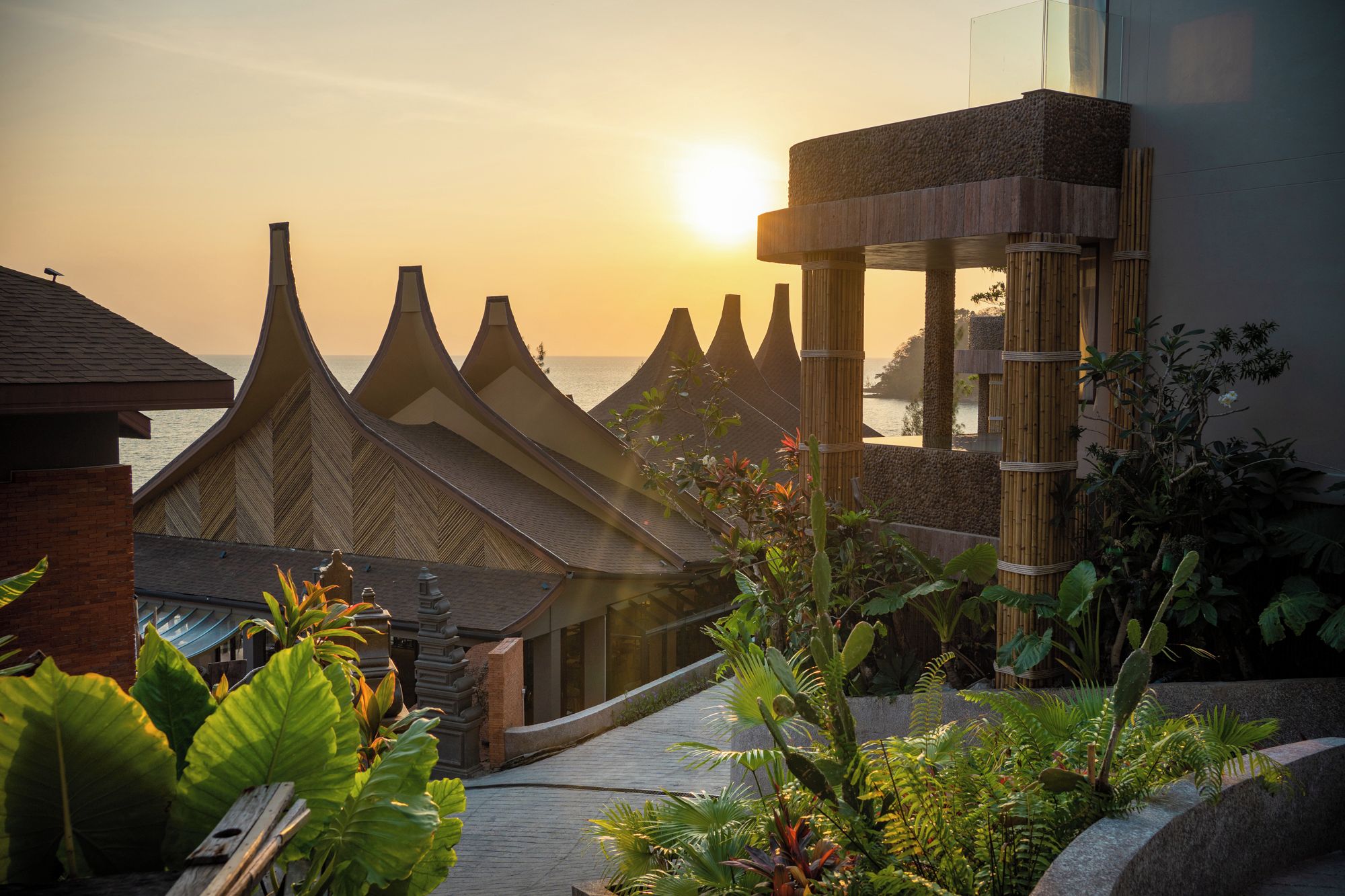 A traditional building with pointed roofs, a bamboo structure, and a view of the sea at sunset.