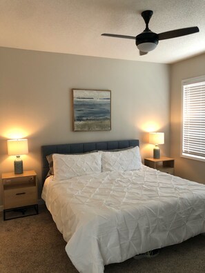 Une chambre à coucher avec un lit, deux tables de chevet avec des lampes, un ventilateur de plafond, un tableau au mur et une fenêtre avec des stores.